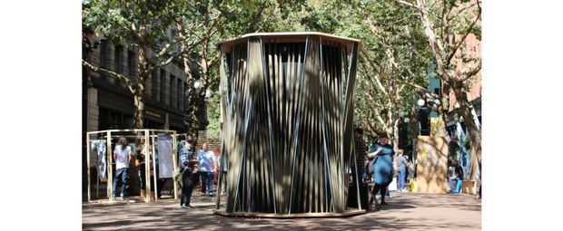 An outdoor architectural installation or pavilion in a leafy public setting. The compact cylindrical structure has a rounded roof and a striking facade of closely spaced, irregular vertical timber-like slats, with people gathered around it and additional temporary display structures in the background.