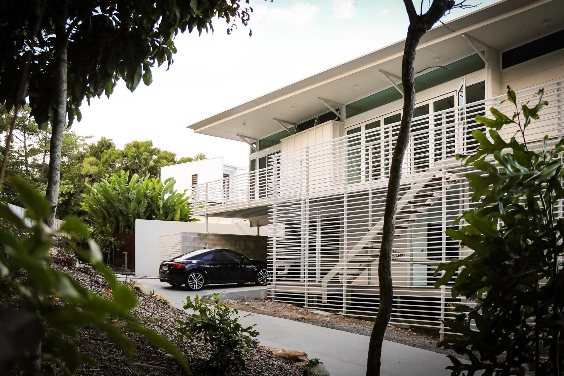 105 Residence - Exterior. Slatted white screens wrapping the upper deck above a leafy garden.