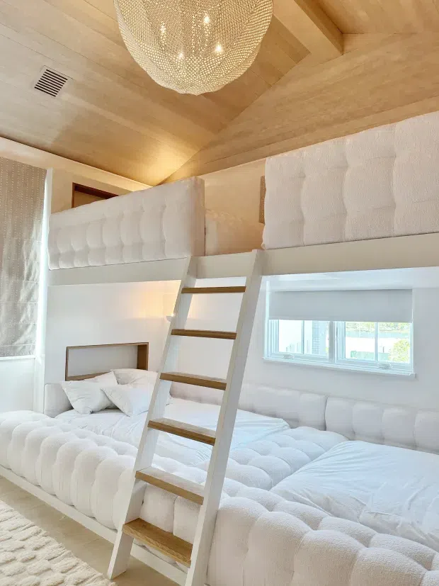 Bright bunk bedroom with two levels of plush white beds, a central white ladder with timber treads, and softly textured upholstered bed surrounds. The room features extensive light timber panelling across the ceiling and upper walls, a large woven pendant light, white roller blinds, and abundant white bedding and upholstery.