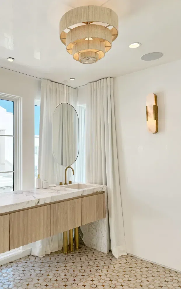 A refined bathroom vanity area with a floating light timber vanity, white marble benchtop and integrated basin, brushed brass tapware and exposed plumbing. An oval mirror hangs in front of full-height white curtains beside tall windows. The room features patterned geometric floor tiles, glossy white walls, recessed ceiling lights, a sculptural tiered woven pendant, and a decorative wall sconce.