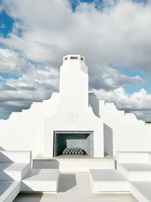 A bright white stucco rooftop or terrace space dominated by a sculptural built-in fireplace and tall stepped chimney structure. The symmetrical architectural composition features curved and stepped parapet forms, integrated white seating platforms, and a dark tiled fireplace surround containing rounded decorative elements. The scene is photographed outdoors beneath a dramatic partly cloudy sky.