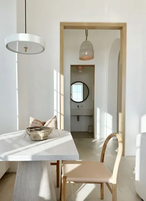 A bright, refined dining area with a pale wood rectangular dining table, curved-back wooden chair, woven centerpiece, and a large white pendant light. A timber-framed doorway opens directly to a compact bathroom featuring vertically ribbed wall treatment, a wall-mounted basin, round mirror, and pendant light. Strong sunlight creates geometric shadows across the white walls and pale flooring, while a second arched doorway and mirror extend the view through the interior.