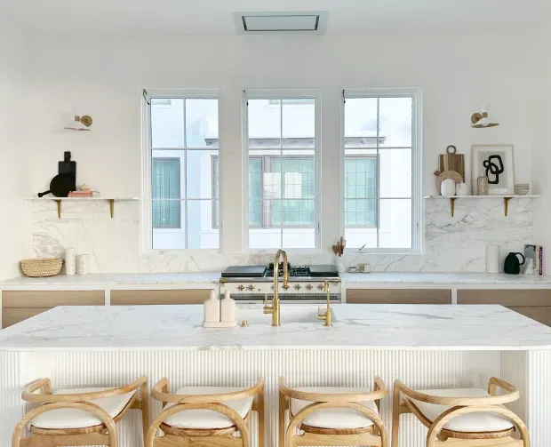 Bright, refined kitchen interior centred on a large marble-look island with a fluted white base and four light timber bar stools. The room features white cabinetry, a marble-look backsplash and benchtops, brass tapware and hardware, open timber shelves with restrained decor, and three tall windows providing abundant natural light.