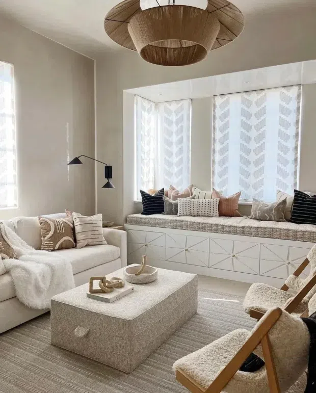 A bright, neutral-toned living room with a large upholstered sofa, built-in window seat, textured ottoman coffee table, lounge chair, patterned cushions, and a sculptural woven pendant light. The room features a geometric white built-in window-seat cabinet, sheer patterned curtains, and soft layered textiles, creating a refined and cozy contemporary setting.