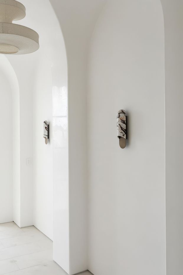 A bright minimalist interior hallway or entry passage with smooth white walls, rounded arched openings, pale tiled flooring, and sculptural wall sconces. A layered woven pendant light is partially visible at the upper left, while daylight enters through a narrow opening or window. The space has a clean, serene, softly curved architectural character.