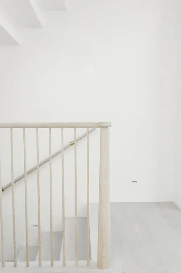 Minimalist bright interior view of a stair landing, featuring a pale timber stair balustrade with slender vertical spindles, a rounded newel post, and a diagonal handrail. White walls, light flooring, and simple geometric ceiling details create an airy, understated space.