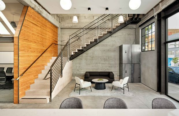 Open-plan commercial or studio interior featuring a prominent exposed concrete stair and mezzanine structure, black metal balustrades, a warm timber-clad stair wall, polished concrete flooring, industrial pendant lighting, and a small seating area beside tall black-framed windows. The space has an industrial warehouse-like character with visible structural services and a compact meeting or lounge zone.