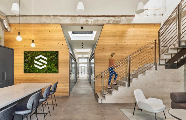 Bright open-plan commercial or industrial-style interior with a central corridor, exposed concrete beam and polished concrete floor. Warm horizontal timber wall panelling contrasts with dark cabinetry and metal-framed glazing. A concrete stair with slim metal balustrade rises to the right, while a long communal island or work counter with stools occupies the left. Pendant lights, skylights and a green wall-mounted display add visual interest; lounge seating is visible in the foreground.