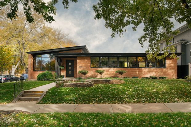 Street-facing view of a low, single-storey brick building with a long band of dark-framed windows, a shallow sloping roof, and a recessed glazed entry. Mature trees and a grassy verge surround the property, with a concrete path and steps leading to the entrance. The facade has a restrained mid-century-inspired composition with strong horizontal lines and extensive glazing.