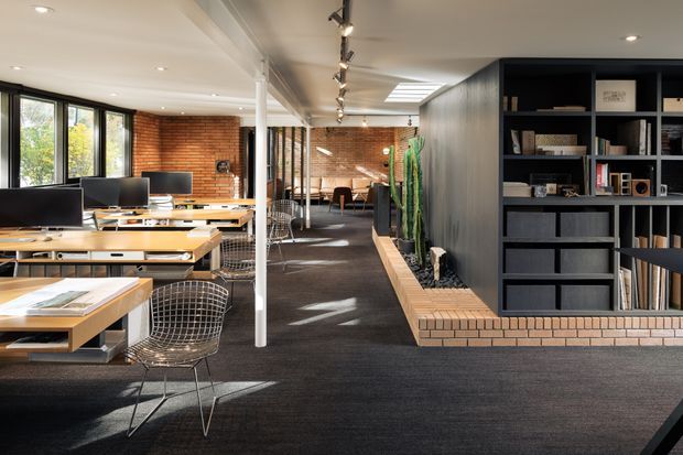 Open-plan contemporary office interior with multiple timber workstations and computer monitors on the left, a central circulation aisle, exposed red-brick walls, black built-in shelving and storage, and an integrated brick planter bench with greenery. The space has a white ceiling with recessed and track lighting, skylights, extensive black-framed glazing, and dark carpet flooring.