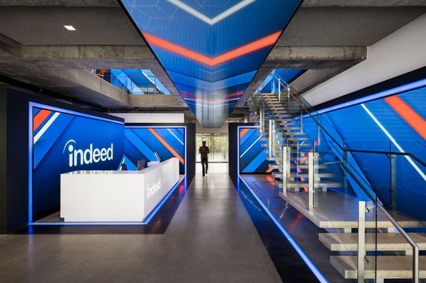 A dramatic modern commercial office reception and circulation space featuring a white reception desk with Indeed branding, vivid blue and orange graphic wall panels, exposed concrete ceilings and beams, polished concrete flooring, and an open steel-and-glass staircase. A bold illuminated blue ceiling feature continues through the space, creating a high-impact branded workplace interior.