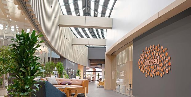 A bright, contemporary commercial interior with a spacious reception or lounge area. The space features a high glazed barrel-vaulted skylight, sweeping curved timber-lined wall, exposed vertical slat detailing, integrated timber-framed glazing, indoor planting, and casual tan lounge seating. A prominent circular timber wall logo feature is visible on the right.