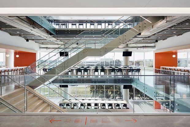 A bright, open-plan commercial interior centered on a broad glass-and-metal staircase spanning multiple levels. Exposed services and ductwork run across the ceiling, while extensive glazing overlooks the surrounding area. Dining or breakout seating is visible on the levels, with vivid orange-red feature walls and transparent glass balustrades.