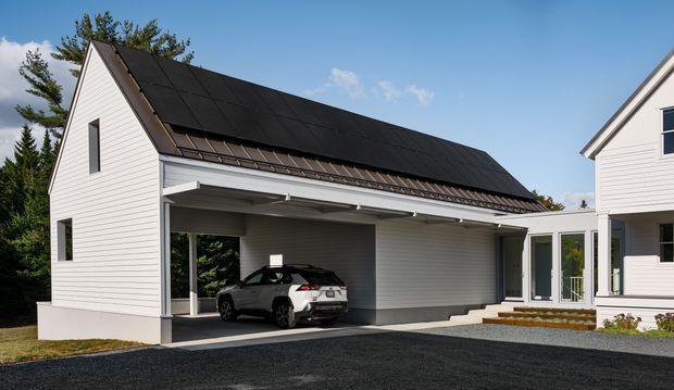 Exterior view of a contemporary white weatherboard-clad residential property with a steep gabled roof covered in dark solar panels. The main structure forms a large open-sided covered carport or garage bay, with a parked vehicle, recessed downlighting, and a glazed connecting passage to a second building. The setting includes paved drive surfaces, trees, and a clear sky.