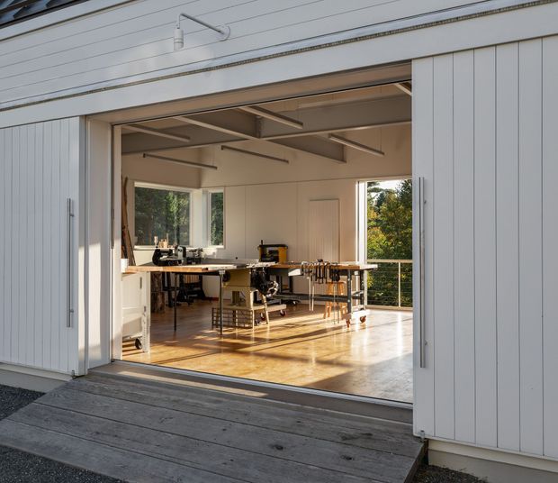 View from a timber deck into a bright, open workshop or studio with fully opened white vertical-panel doors. The space has polished timber flooring, large windows, exposed structural ceiling beams, workbenches, stools, tools, and craft equipment, with greenery visible outside.