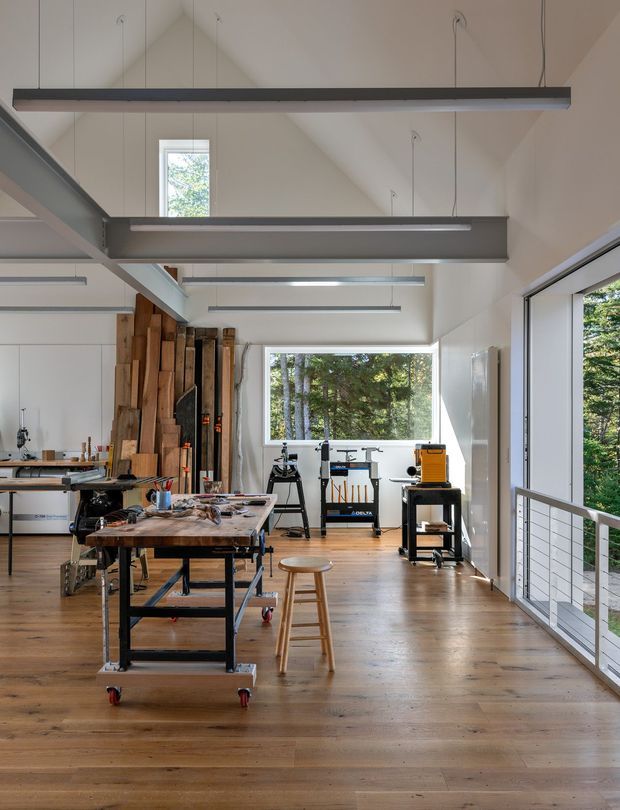 A bright, open-plan woodworking workshop or creative studio with a high pitched ceiling and exposed structural beams. Large windows and glazed doors bring in views of trees, while linear suspended strip lights illuminate timber workbenches, rolling tool tables, woodworking equipment, and vertically stored timber boards. The space has warm timber flooring, white walls, dark metal detailing, and a balcony-like metal cable railing along the glazed edge.