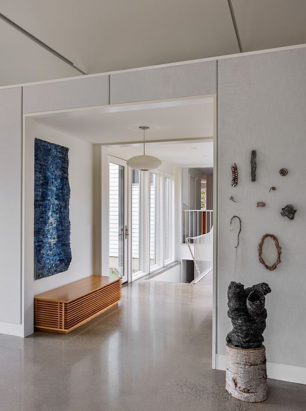 A refined contemporary entry hall with pale textured built-in wall panels, a polished concrete floor, and a timber slatted console. The hallway opens toward a glazed circulation zone and stairway, with a sculptural pendant, blue textured wall art, and curated sculptural objects displayed on the wall and plinth.