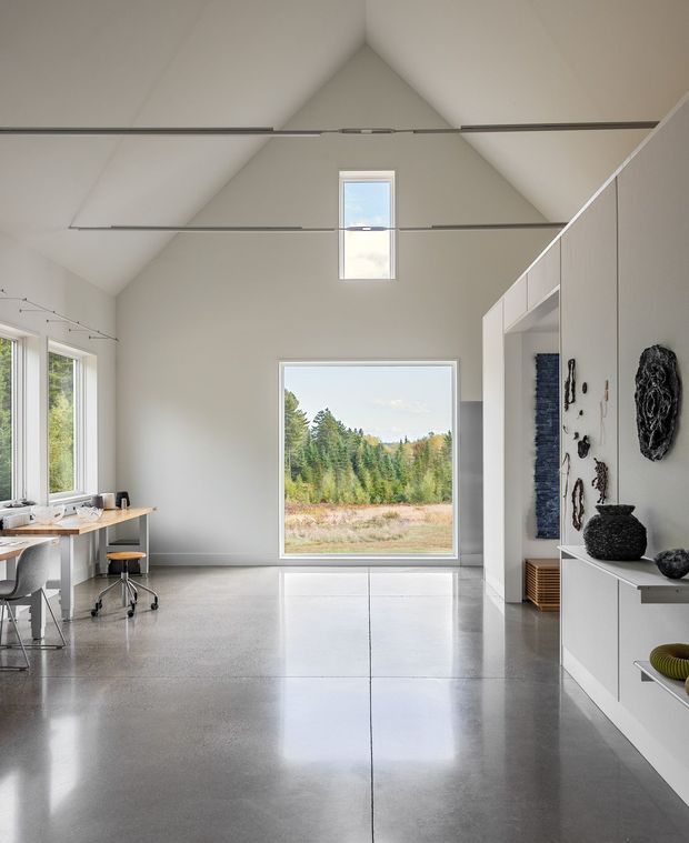 A bright, minimalist open-plan interior with a dramatic vaulted ceiling, polished concrete floor, and large windows framing a natural landscape. A compact desk workspace sits beside the windows, while sleek white built-in cabinetry and open shelving line the right wall, displaying dark sculptural objects. A high-level skylight and suspended linear track elements emphasize the room's height and clean geometry.