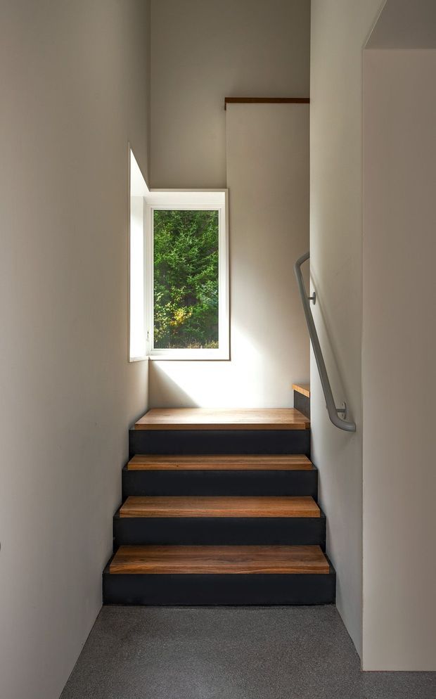 A narrow, minimalist stairway with dark painted stair risers and warm timber treads, rising toward a small white-framed window with leafy greenery outside. Neutral plaster walls, grey carpet at the lower landing, and a simple grey metal handrail create a restrained, contemporary interior.
