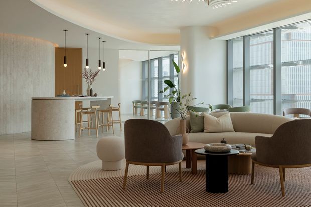 A spacious, refined open-plan apartment interior combining a kitchen, dining area and living room. The room features floor-to-ceiling glazing, pale stone-look tiled flooring, a curved light-toned kitchen island, timber bar stools, sculptural pendant lighting, curved upholstered lounge chairs, a cream sofa, a textured neutral rug and abundant indoor planting. The palette is warm and softly neutral with contemporary furnishings and strong natural light.