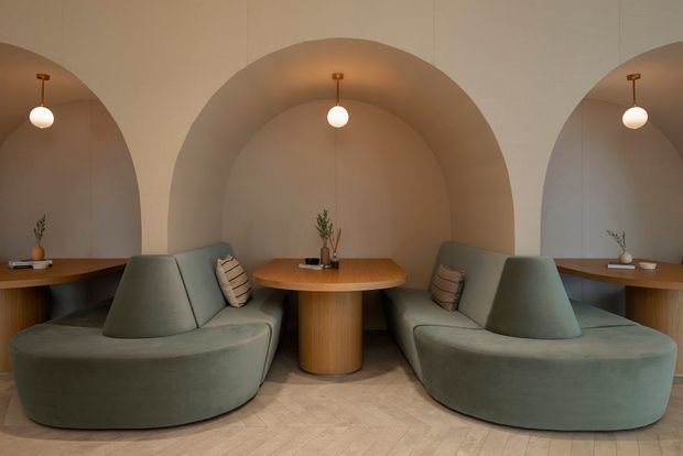 A refined interior seating area with three repeated arched alcoves, each containing a small round timber table and pendant light. Curved upholstered booth seating in muted sage-grey wraps around the central tables, with cylindrical timber pedestal bases and small decorative plants. The space has softly plastered walls and a pale geometric tiled floor, creating a calm, sculptural, boutique hospitality-like atmosphere.