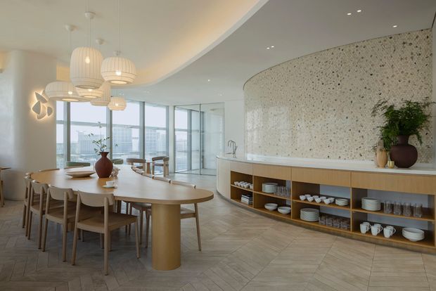 A refined open-plan dining and kitchen space with a long rounded dining table, timber dining chairs, sculptural pendant lights, and a curved kitchen island with open timber shelving. The kitchen features a pale terrazzo-like curved wall and benchtop, light timber joinery, extensive floor-to-ceiling glazing, and a herringbone-patterned floor. The overall aesthetic is bright, warm, contemporary, and softly organic.