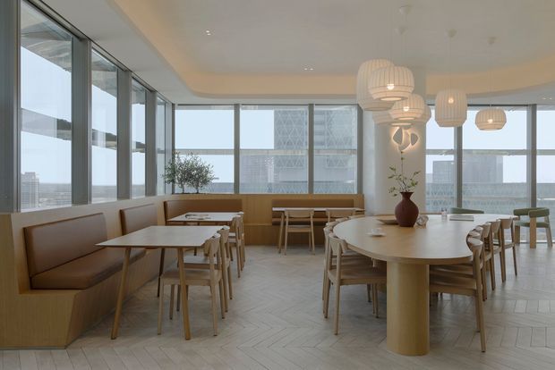 Bright contemporary dining and lounge area with extensive floor-to-ceiling glazing, pale herringbone flooring, integrated tan upholstered banquette seating, light timber tables and chairs, and sculptural pendant lights. The space overlooks an urban waterfront or coastal setting, with a large structural column and decorative planting providing focal points.