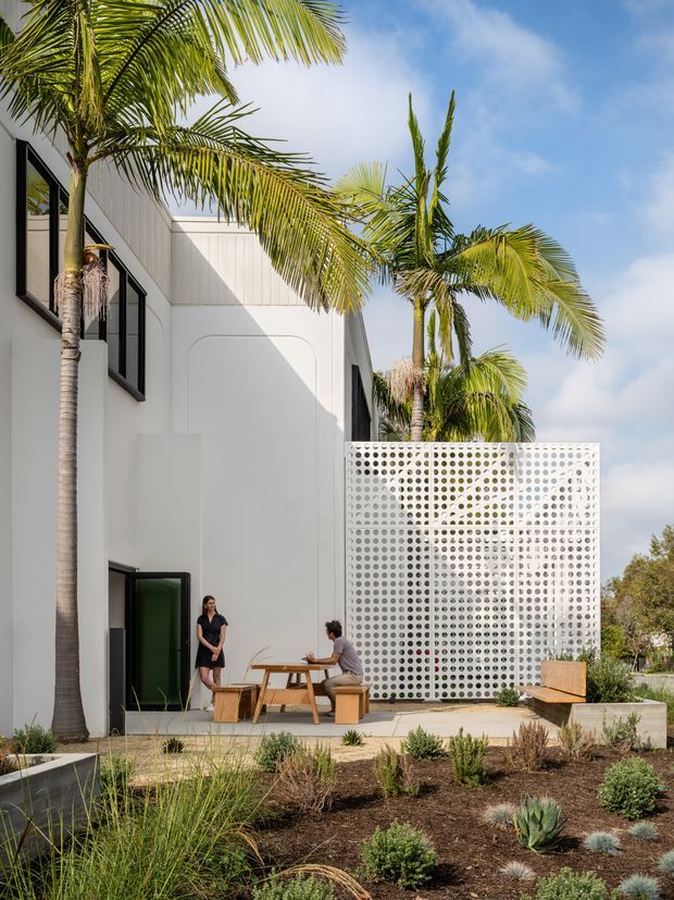 A bright contemporary white multi-storey building exterior with black-framed windows, vertical wall cladding, and a distinctive perforated breeze-block privacy screen enclosing a small outdoor dining area. Tall palm trees and drought-tolerant landscaped planting surround the paved courtyard, creating a tropical yet minimalist setting.