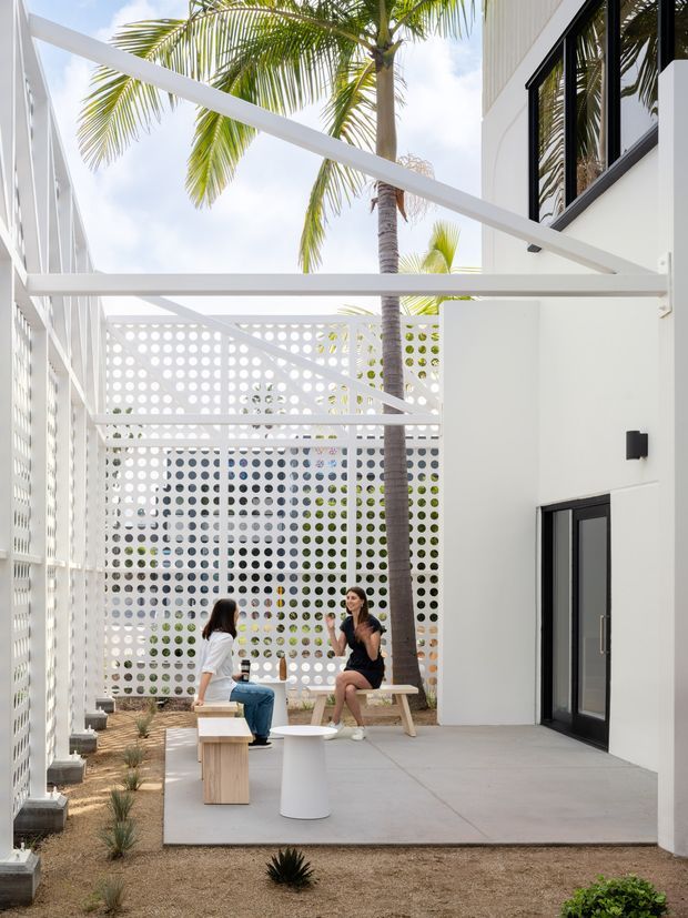 Bright contemporary courtyard beside a white building, featuring a white pergola-like overhead structure, perforated breeze-block privacy screens, black-framed glazing and doors, a concrete-paved seating area, and tropical palm landscaping. Two people sit on a timber picnic-style bench with simple outdoor stools and tables. The composition highlights the architectural courtyard enclosure and filtered light.