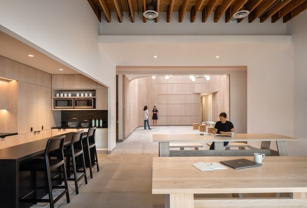 Open-plan contemporary interior combining a kitchen, dining/work table and spacious lounge or communal area. The kitchen features full-height light timber cabinetry, a dark benchtop, integrated appliances and a long island with black bar stools. Light timber tables occupy the foreground, while the rear space has vertically ribbed timber wall panelling and a large open ceiling volume. Exposed timber ceiling joists with recessed and circular downlights add strong architectural character. The palette is warm and neutral with timber, beige, white and charcoal accents.