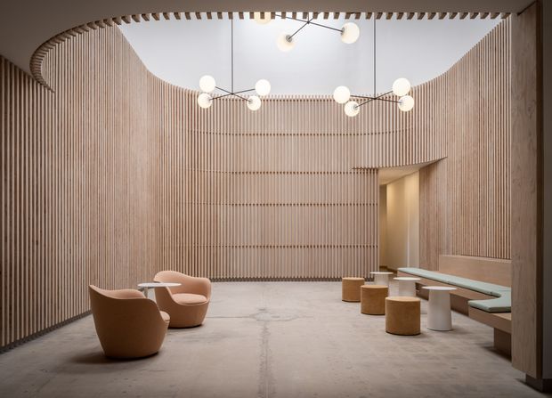 A spacious, softly lit contemporary lobby or lounge with curved walls lined in vertical timber battens, a pale polished concrete floor, sculptural globe pendant lighting, and minimalist seating including upholstered lounge chairs, small stools, and a built-in bench. The room has a high, partially open ceiling and a refined neutral palette.