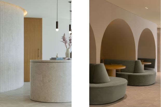 A split architectural interior image showing a softly minimalist hospitality or commercial space. On the left is a rounded textured plaster feature wall, timber door, cylindrical pale stone or terrazzo reception counter, and suspended globe pendant lights. On the right are sculptural curved upholstered lounge seats arranged around small round timber tables within repeated large arched wall niches. The palette is warm neutral with beige, taupe, pale timber, and muted olive-grey upholstery.