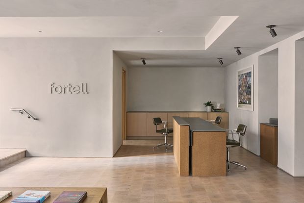 A minimalist contemporary commercial interior, likely a reception or showroom, with pale stone or tile flooring, smooth white walls, warm timber cabinetry, a central wood-clad island or counter, and black track lighting. Several modern office-style chairs and a small desk area occupy the rear, while a raised wall logo and framed artwork add subtle branding and visual interest.