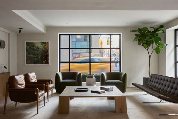 A refined contemporary living room with a low, pale ceiling, large black-framed grid windows, and a neutral palette. The room contains two dark green curved lounge chairs, a tufted dark leather sofa, leather armchairs, and a substantial rectangular timber coffee table. A tall indoor tree and framed artwork add warmth, while the urban street scene is visible through the glazing.
