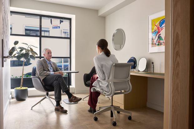 A bright, modern consultation or work room with two people seated across from each other. The space features a large black-framed window, pale walls, a light timber floor, a built-in timber desk with a mirror and small tabletop objects, contemporary office chairs, a potted plant, and colourful framed artwork. A timber-framed opening is visible at the right edge.
