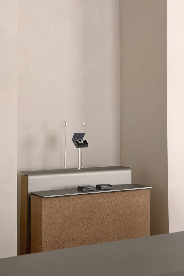 Minimalist bathroom vanity viewed straight on, featuring a sculptural integrated basin or backsplash form, wall-mounted chrome tapware, and a dark slim benchtop over a textured cork-toned vanity cabinet. The space has tall warm off-white plaster walls and a restrained, contemporary product-photography aesthetic.