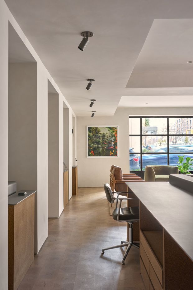 A contemporary open-plan interior viewed along a kitchen or dining zone, with a long dark stone island and integrated cabinetry on the right, stools, timber flooring, warm neutral plaster walls, recessed ceiling planes, and track spotlights. A corridor with built-in joinery leads toward a bright living area featuring lounge seating and large black-framed windows. The space has a refined, minimalist fit-out with natural light and muted earthy tones.
