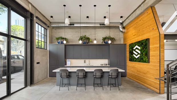 A contemporary industrial-style commercial kitchen and breakout area with a long island bench and four stools. The space features exposed concrete walls and ceiling services, black-framed floor-to-ceiling glazing, dark grey full-height cabinetry, a white stone-look benchtop, pendant lighting, and a warm timber-lined feature wall with a planted logo panel. A stairway is visible at right.