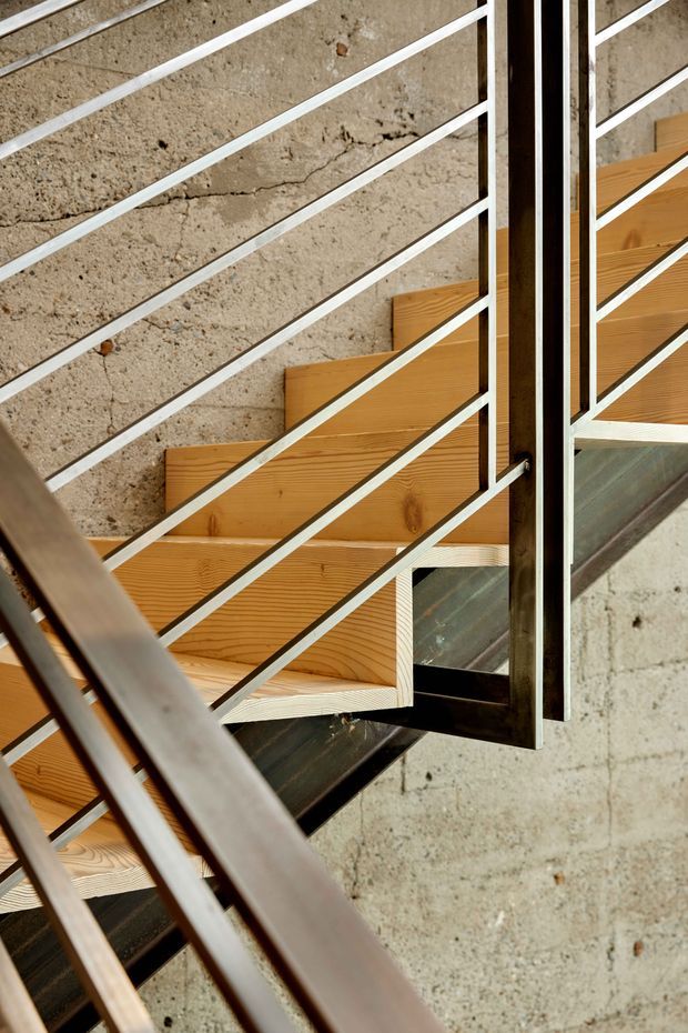 Close-up architectural view of a modern staircase with warm timber treads and stringer elements, set against a rough exposed concrete wall. Slim white-painted metal balustrade rails create strong diagonal lines, while dark metal structural components and a timber-framed glazed opening add contrast. The composition emphasizes the stair construction, material textures, and geometric detailing.