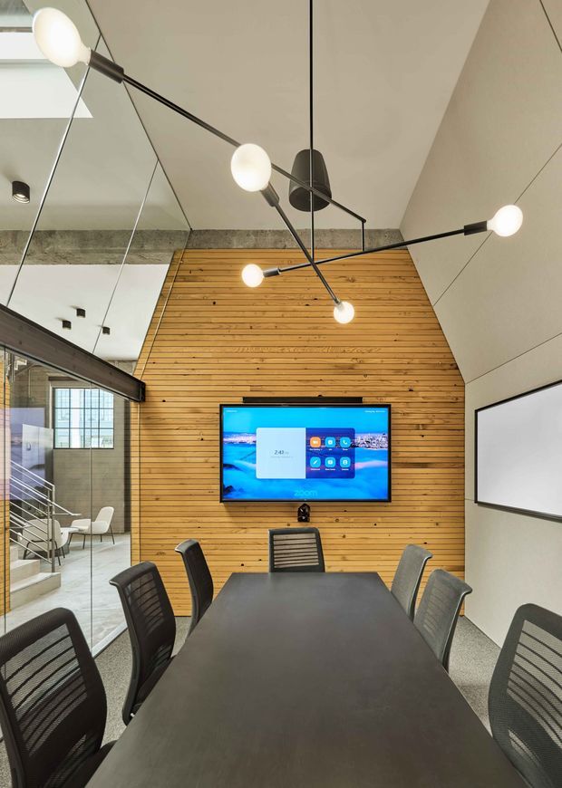 A contemporary commercial meeting room with a long dark conference table surrounded by mesh office chairs. The room features a dramatic multi-arm globe pendant light, a warm horizontal timber feature wall with a wall-mounted display, a large white presentation screen, exposed concrete beam, and glass partitioning revealing an adjacent stair and workspace.