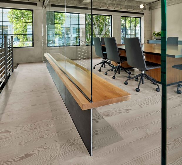 A modern open-plan office interior with a long timber conference table and dark upholstered swivel chairs, enclosed partly by clear glass partitions. The space features exposed concrete ceilings and columns, black-framed industrial-style windows, pale timber flooring, and a minimalist, utilitarian fit-out.
