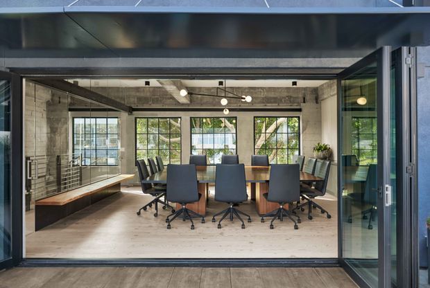 A modern industrial-style meeting room viewed through wide-open glazed folding or sliding doors. The space features a long timber conference table surrounded by dark office chairs, exposed concrete walls and ceiling, black-framed grid windows, linear built-in timber bench seating, and track-style pendant lighting. The room opens directly onto a timber-floored exterior threshold or terrace.