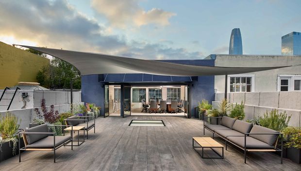 A contemporary rooftop terrace or courtyard entertaining area with expansive grey decking, low-profile outdoor sofas and armchairs, planters, and a large sculptural tensile canopy. The canopy shelters glazed doors opening into an interior dining space, while modern high-rise buildings are visible in the background.