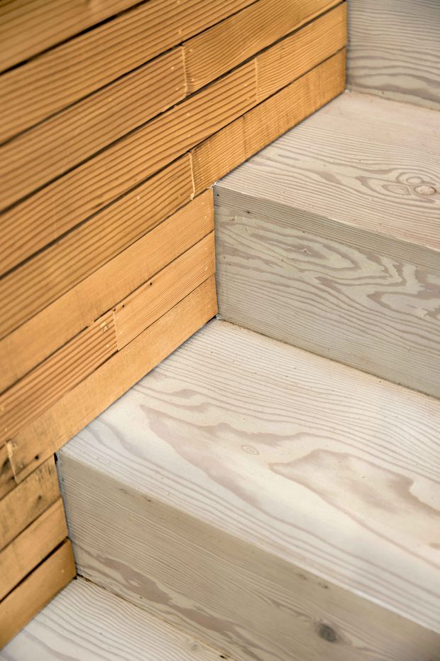 Close-up detail of a timber staircase beside a timber-clad wall. The stair treads and risers show pale, visibly grained wood, while the adjacent wall has warm-toned horizontal slatted timber boards with a textured grooved surface.