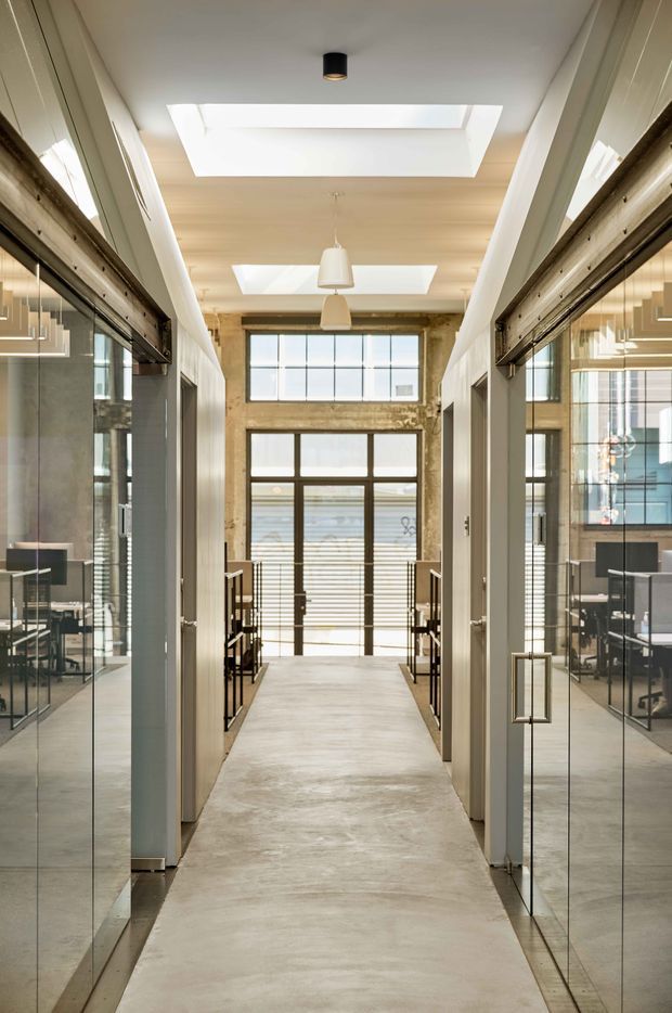 A symmetrical interior corridor in a contemporary commercial workplace, with glass-fronted offices on both sides, exposed warm-toned structural walls at the far end, polished concrete flooring, timber-lined ceiling sections, and large rectangular skylights. Dark metal-framed glazing and doors create a refined industrial character, while pendant lights and abundant daylight brighten the passage.