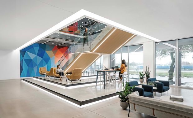 A bright, expansive contemporary office or commercial interior with an open-plan lounge and workspace arranged around a raised platform. The space features floor-to-ceiling glazing, a dramatic angular timber-lined stair or ceiling volume, a mirrored upper-level void, polished light flooring, dark lounge seating, and a large geometric blue, orange, and red feature wall mural. Indoor planting and abundant daylight create an airy, refined atmosphere.