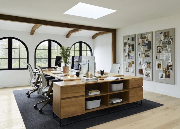 Bright, spacious home office with a long central timber work island, multiple ergonomic office chairs, computers, shelving, and a large pinboard wall. The room has pale timber flooring, white walls, exposed dark timber beams, arched black-framed windows, and a skylight, creating a refined contemporary workspace with traditional architectural accents.