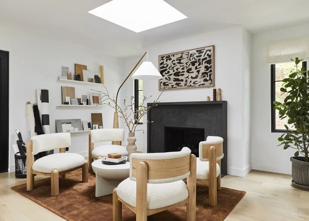 Bright, refined living room with white walls, pale timber flooring, a dark fireplace surround, sculptural cream boucle lounge chairs, a low white coffee table, and a brown area rug. Open shelving displays artwork and objects, while a large abstract artwork hangs above the fireplace. A skylight and tall window provide abundant natural light, complemented by a contemporary floor lamp and indoor plant.