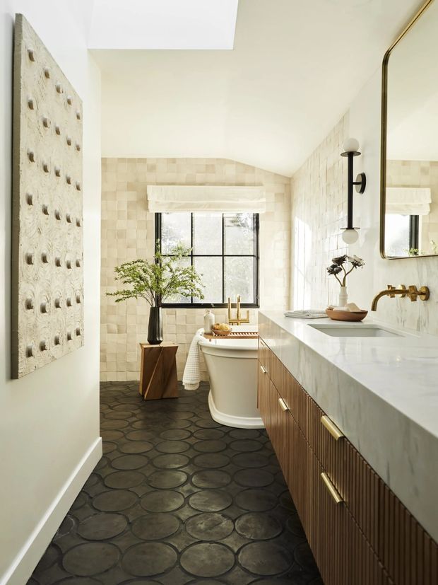Bright, elongated bathroom with a freestanding bathtub beneath a black-framed window, a long fluted timber vanity with a white stone benchtop, twin basins and brass tapware. The walls are clad in softly varied light stone or ceramic tiles, while the floor has a dark circular-patterned finish. A large brass-framed mirror, sculptural wall art, greenery and globe wall lighting add a refined, warm contemporary character.