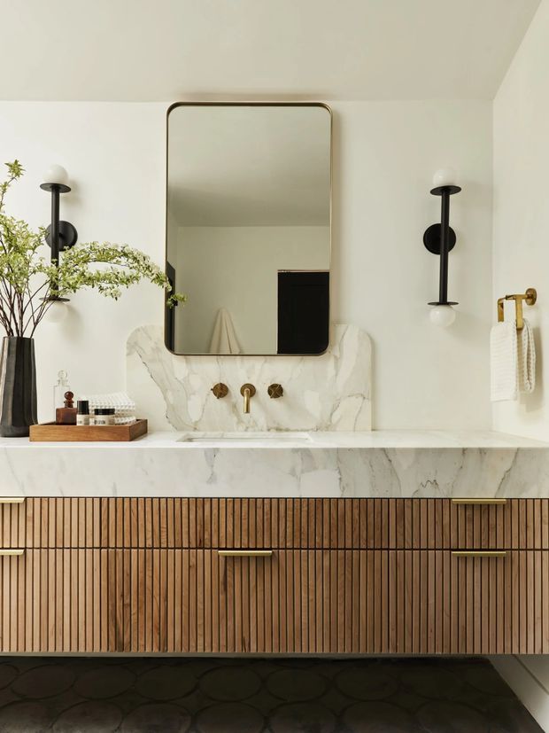 A refined bathroom vanity with a large rounded-rectangle mirror, sculptural black wall sconces, marble backsplash and countertop, brass tapware and towel rail, and fluted timber cabinetry with brass handles. A dark patterned floor and minimal styling create a warm, contemporary look.